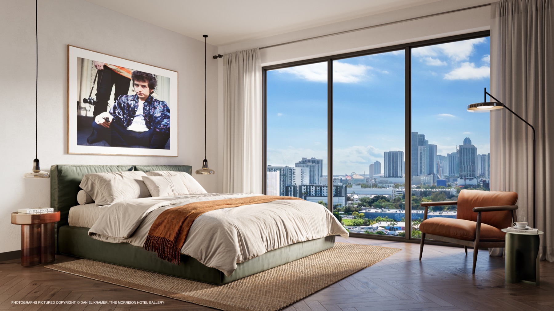 A primary bedroom at The Rider Residences Wynwood — a sage-green upholstered bed, warm wood floors, sculptural pendant lighting, and floor-to-ceiling glass opening to sweeping Miami skyline views.