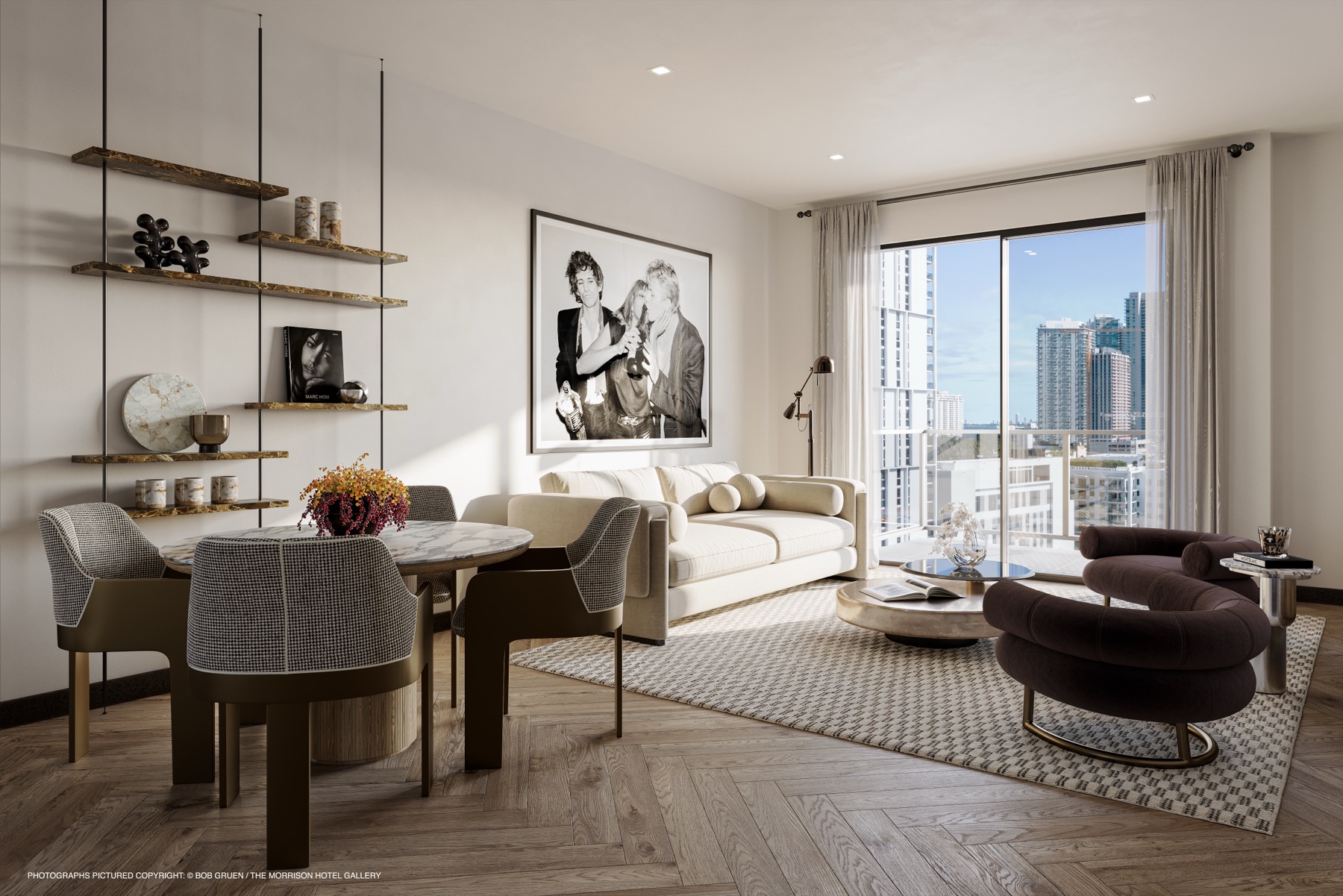 A living room at The Rider Residences Wynwood — herringbone wood floors, a cream sofa and sculptural bronze chairs, floor-to-ceiling glass framing the Miami skyline, and curated black-and-white art. Fully furnished by the developer.